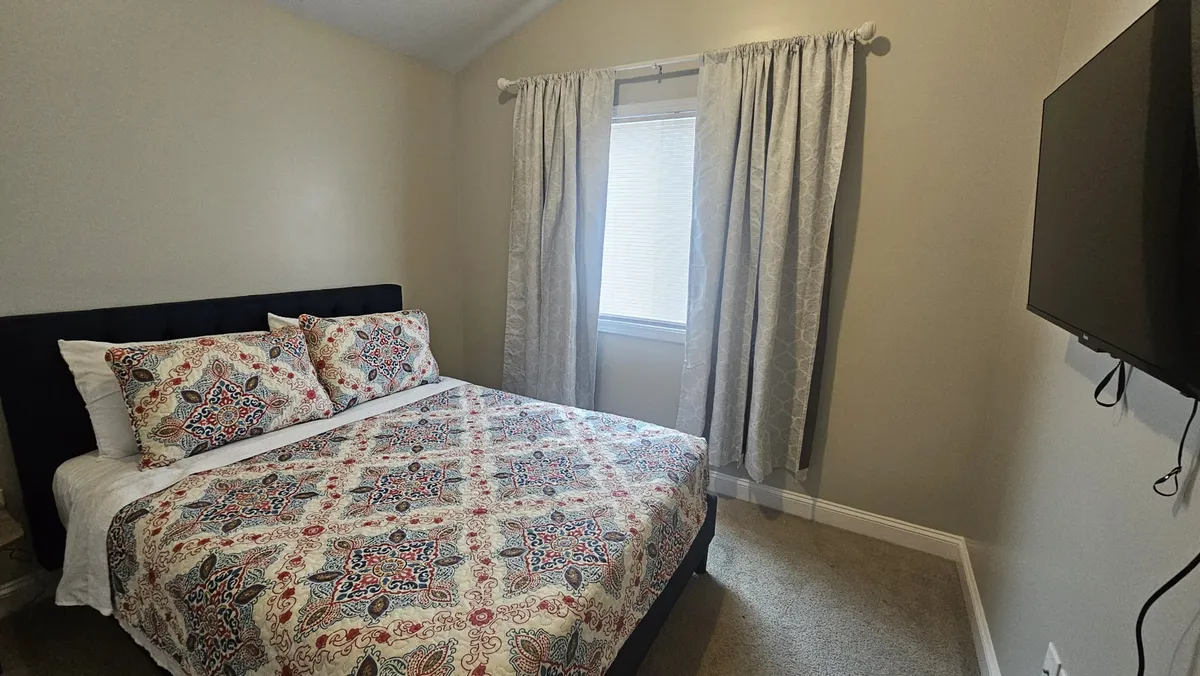 A bedroom featuring patterned bedding, a window with curtains letting in natural light, and a wall-mounted TV—creating a restful environment for winding down.