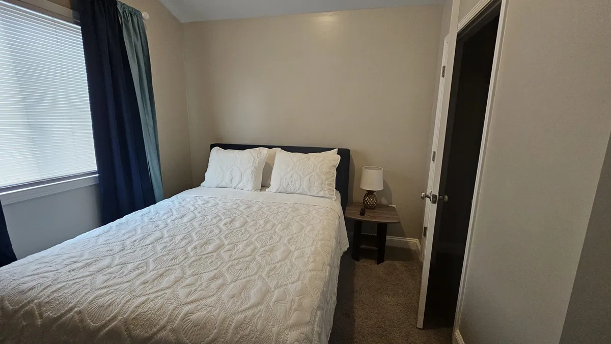 A neatly arranged bedroom with a bed adorned with decorative pillows, a nightstand with a lamp, and a window letting in natural morning light. The setting feels peaceful and inviting, ideal for recuperation.