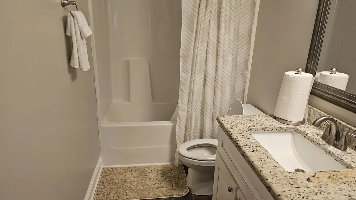A calming bathroom with a granite countertop, large mirror, shower curtain, toilet, and towel rack. The space is designed for relaxation and self-care, offering a refreshing start or end to the day.