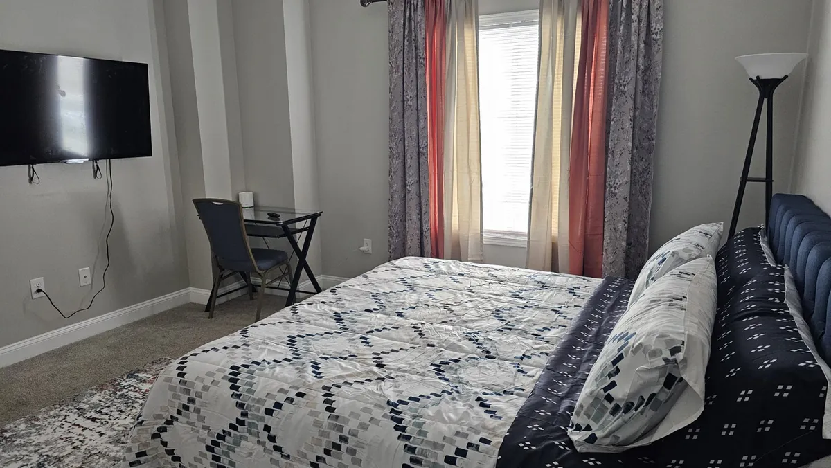 A sunlit bedroom featuring a bed with stylish patterned bedding, a TV mounted on the wall, a window with light filtering through curtains, and a small desk with a chair. The atmosphere is cozy and modern, blending comfort with functionality.