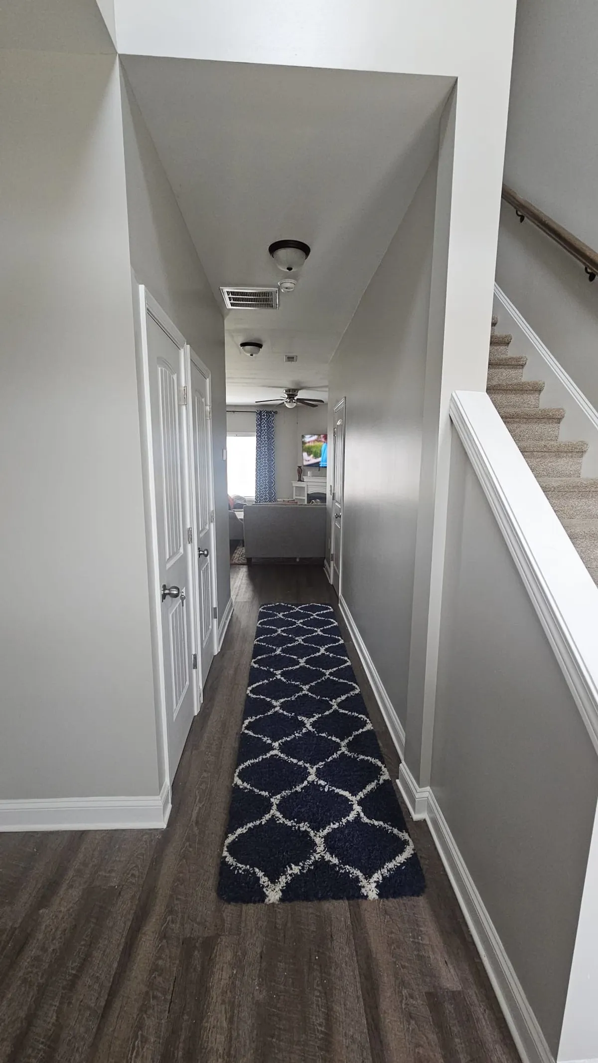 A cozy view looking through a long hallway toward the living room, featuring a textured rug and a staircase, with ambient natural light spilling in. The open transition between spaces hints at an open-plan layout and the welcoming nature of the home.