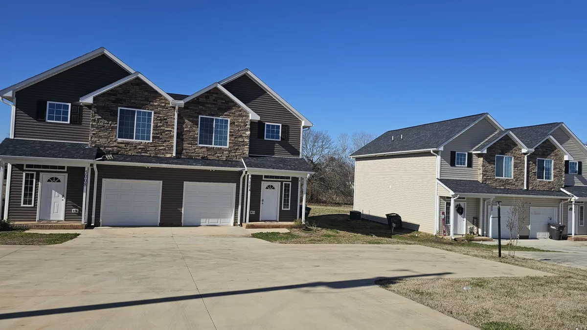 This exterior shot features two duplex buildings side by side, a spacious driveway, and landscaped front yards. The private entrances to each unit are clearly visible, as is the designated parking directly in front of each residence. The overall scene projects a sense of both individual privacy and a welcoming neighborhood atmosphere.