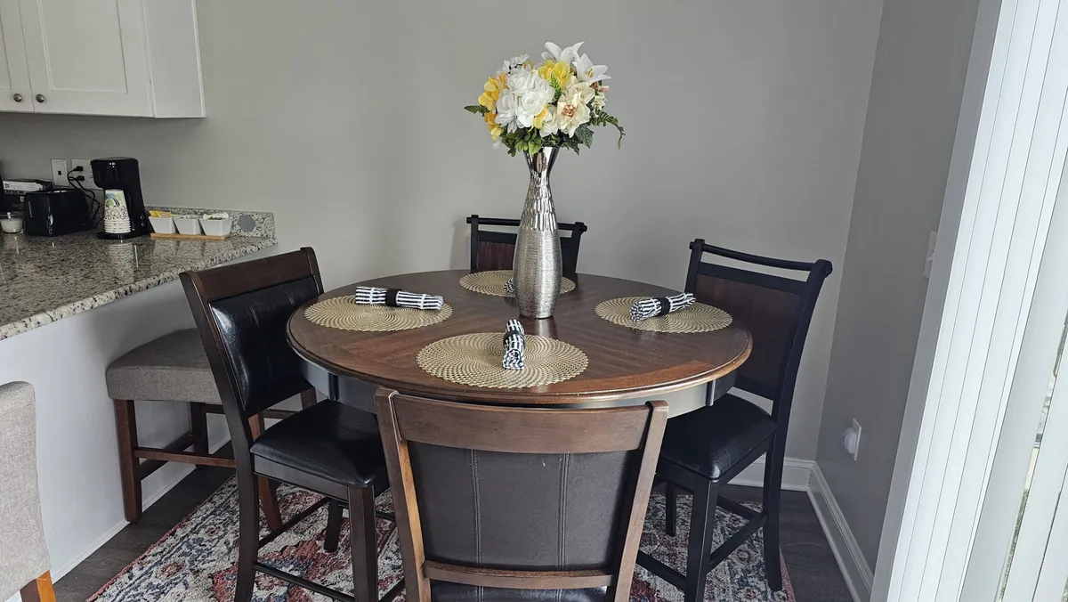 A bright and inviting dining area with a round table set with woven placemats and a floral centerpiece, radiating a welcoming ambiance perfect for shared meals or morning coffee. The setup encourages togetherness and lingering conversations.