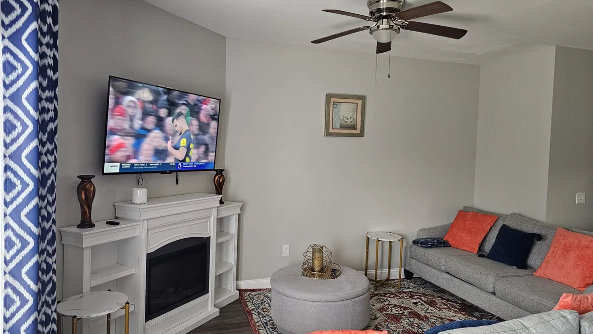 A relaxed living room scene featuring a comfortable gray sofa with colorful accent pillows, a decorative coffee table, and a television displaying a sports game. The layout suggests an open environment ideal for socializing, relaxing with family, or enjoying movie nights.